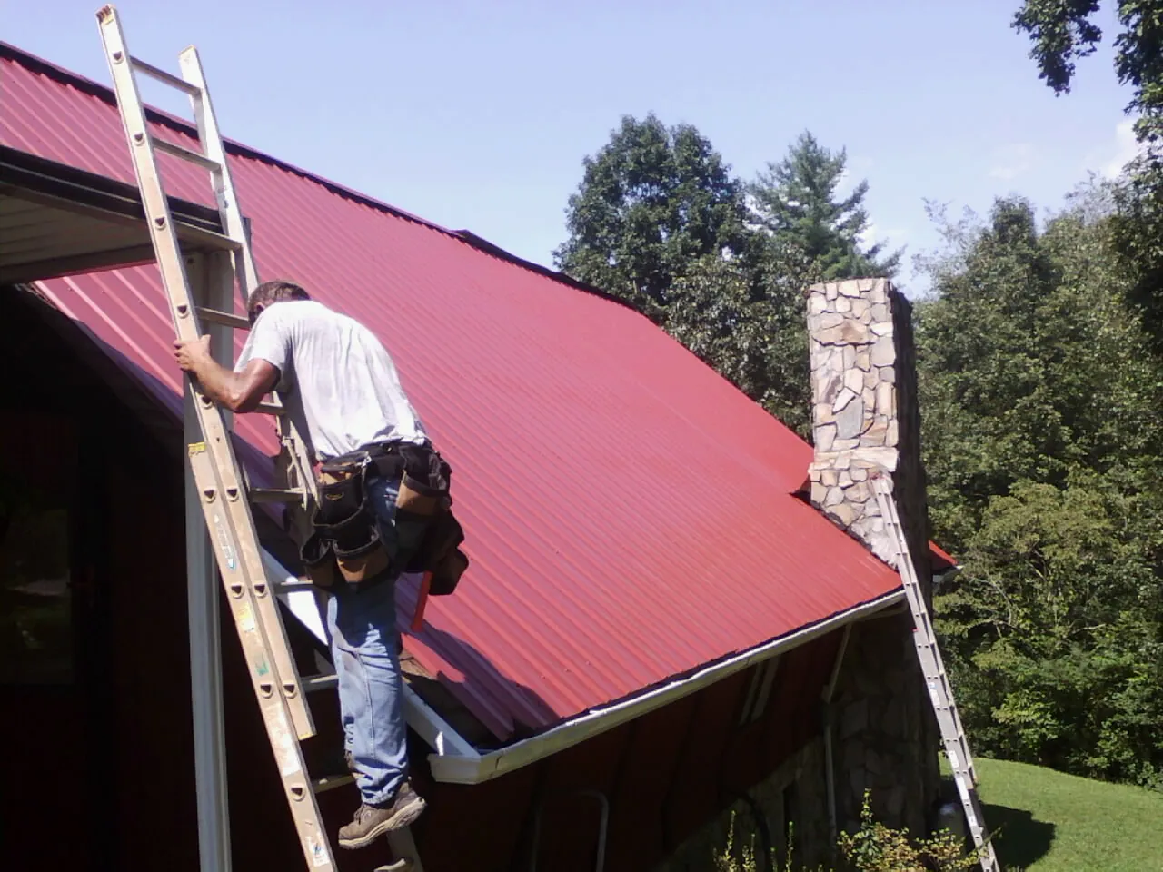 Roofer on a ladder beside a red metal roof and stone chimney — Soffit & Fascia Repair by Roofing Brothers in Crucible